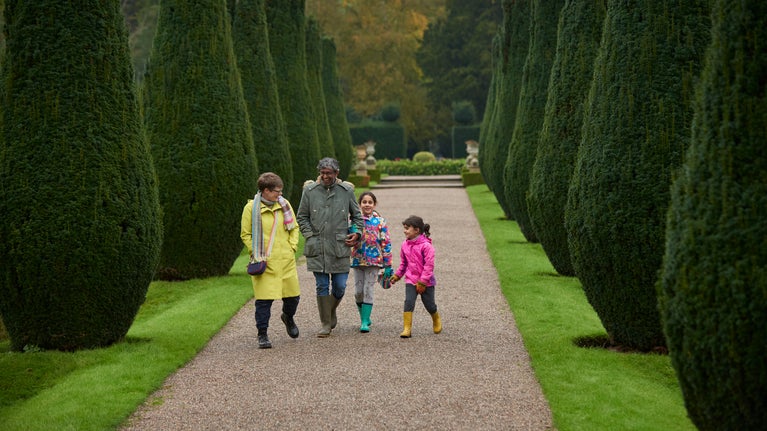 A family consisting of a man, a woman, and two young girls walking down a path in Erddig's garden during autumn. They are smiling and laughing, surrounded by bushes.